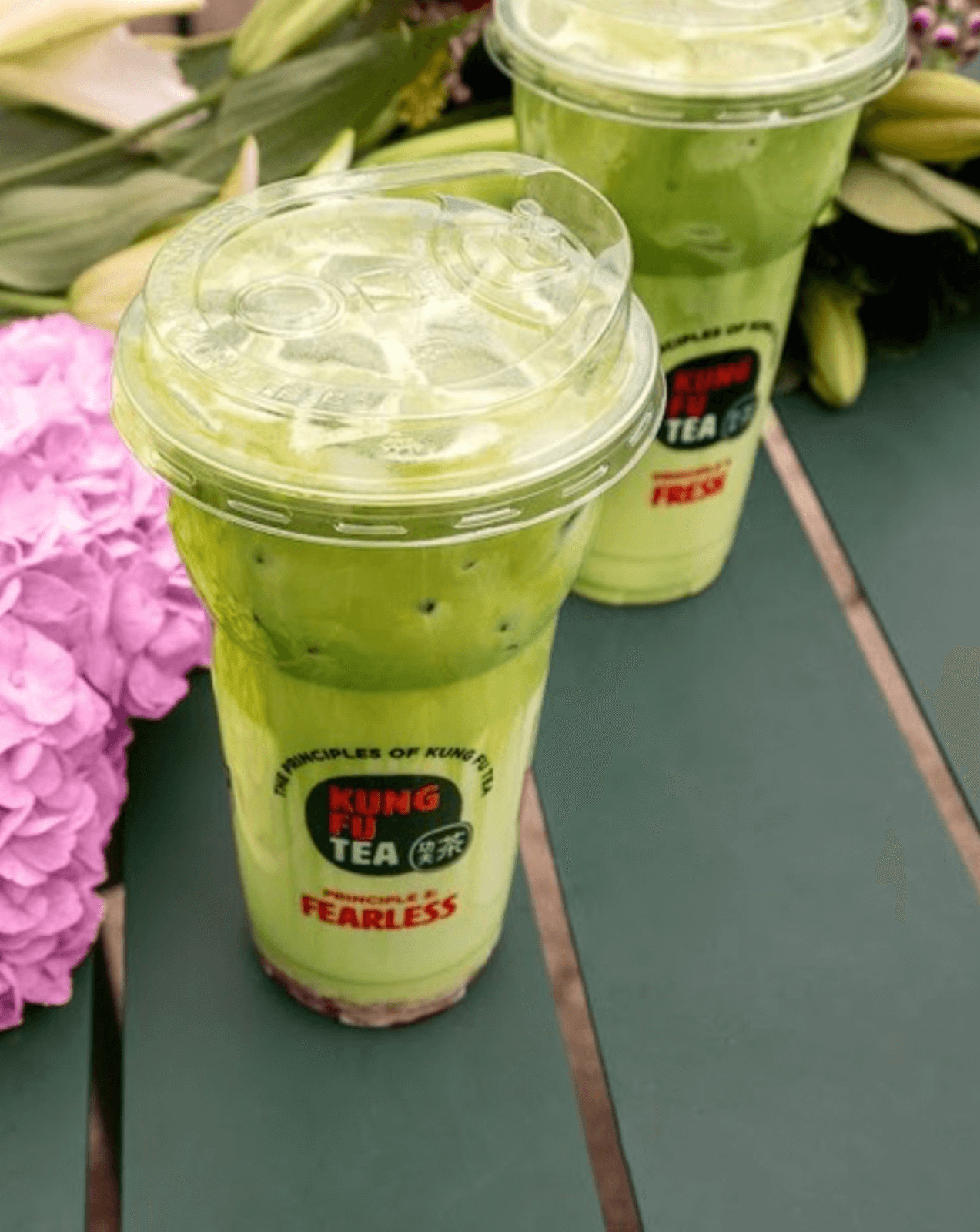 Two plastic cups of green matcha drink from Kung Fu Tea are placed on a green table, highlighting fresh dining concepts among Birmingham restaurants, with purple flowers and foliage in the background.