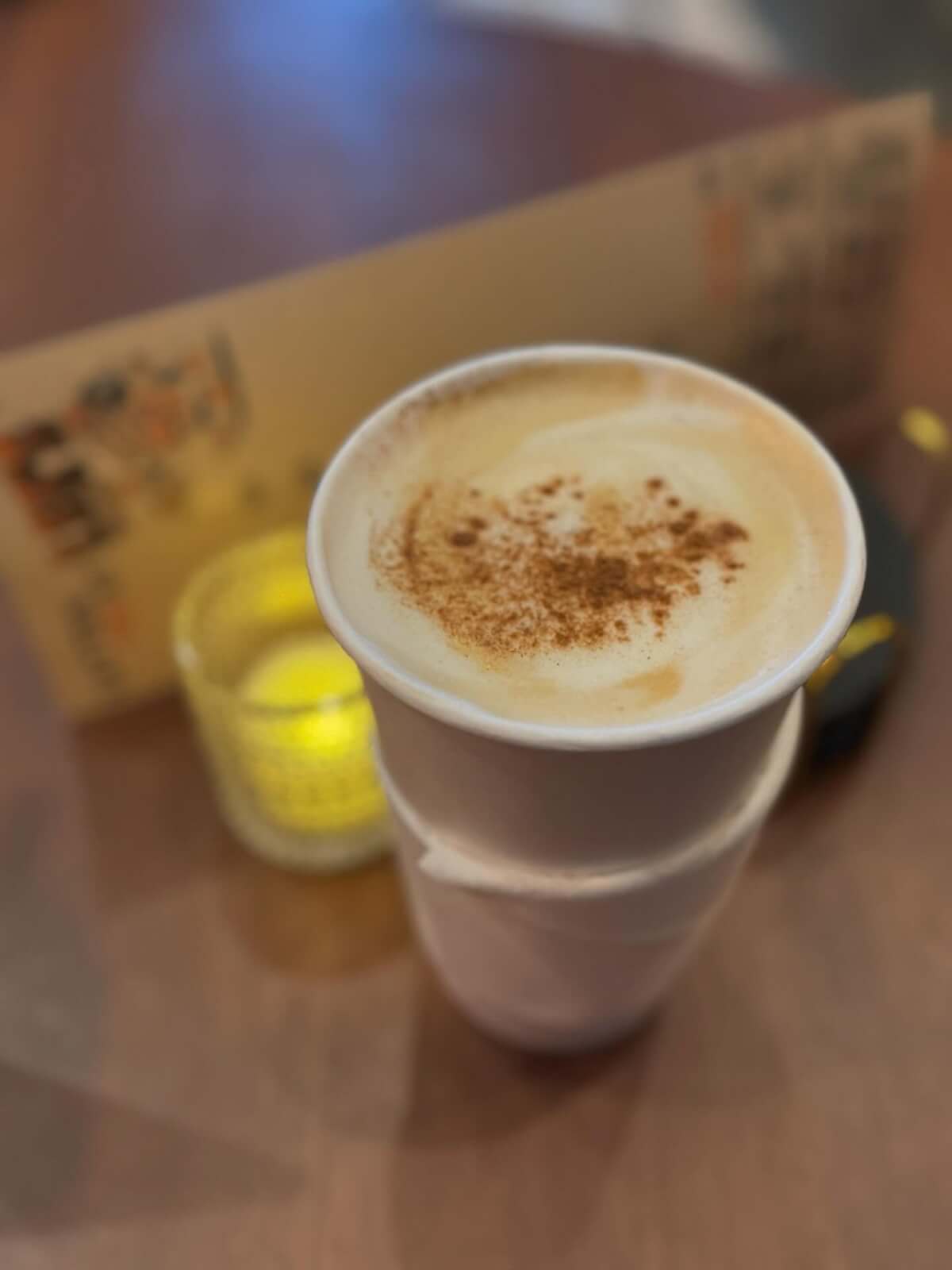 A paper cup of cappuccino with cinnamon on top sits on a wooden table at one of the new restaurants in Birmingham, next to a lit candle and a blurred menu in the background, capturing the cozy spirit of local dining.