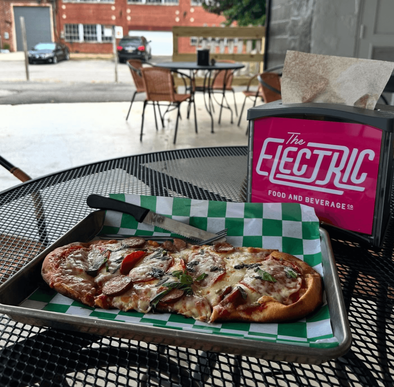 A tray with a flatbread pizza, knife, and fork sits on a black metal table next to a pink napkin dispenser labeled “The Electric Food and Beverage,” one of the standout Birmingham restaurants in this lively outdoor seating area.