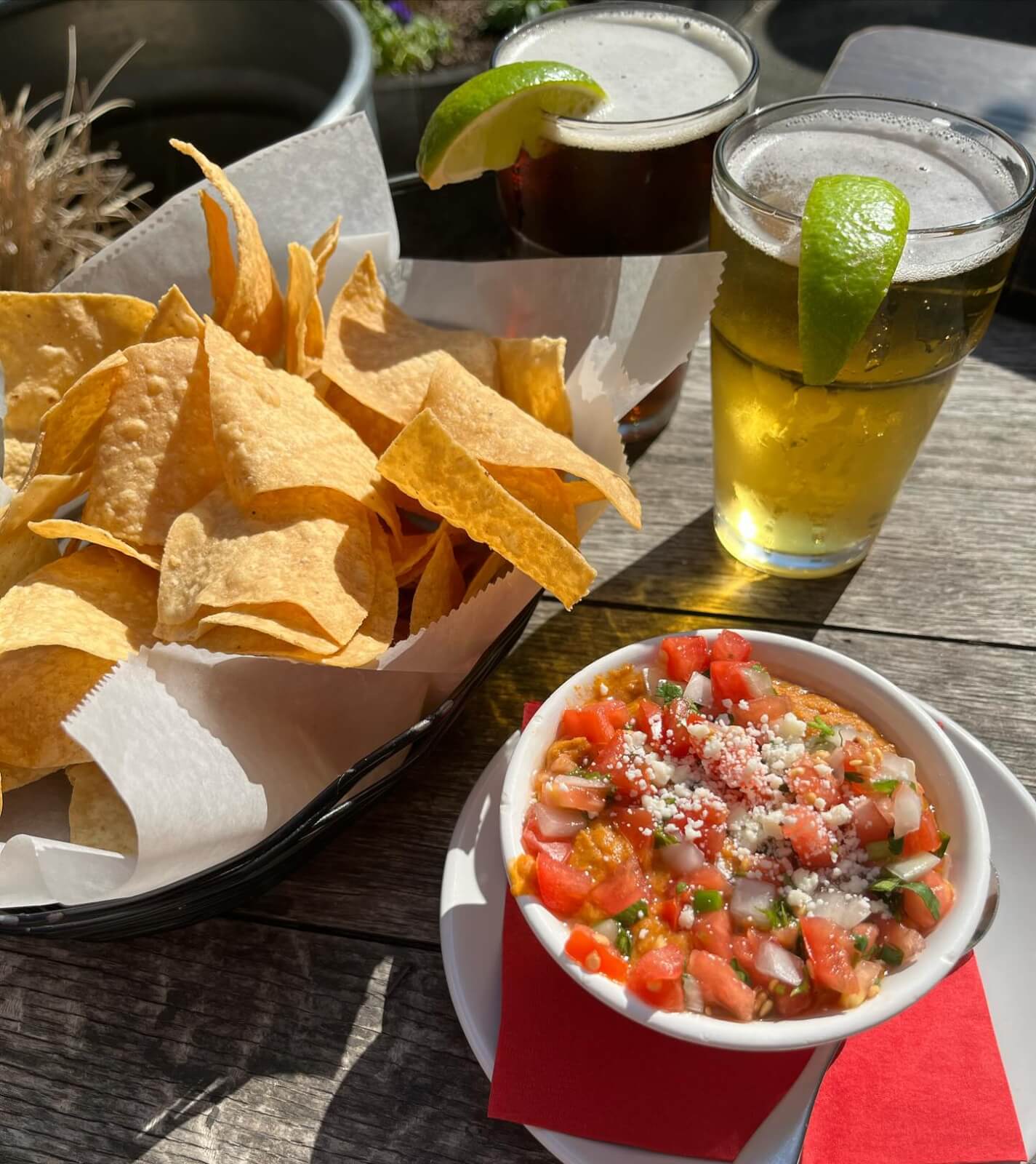 A basket of tortilla chips and a bowl of salsa topped with diced tomatoes and cheese sit next to two glasses of beer with lime wedges on a wooden table—perfect for exploring new restaurants Birmingham has to offer.