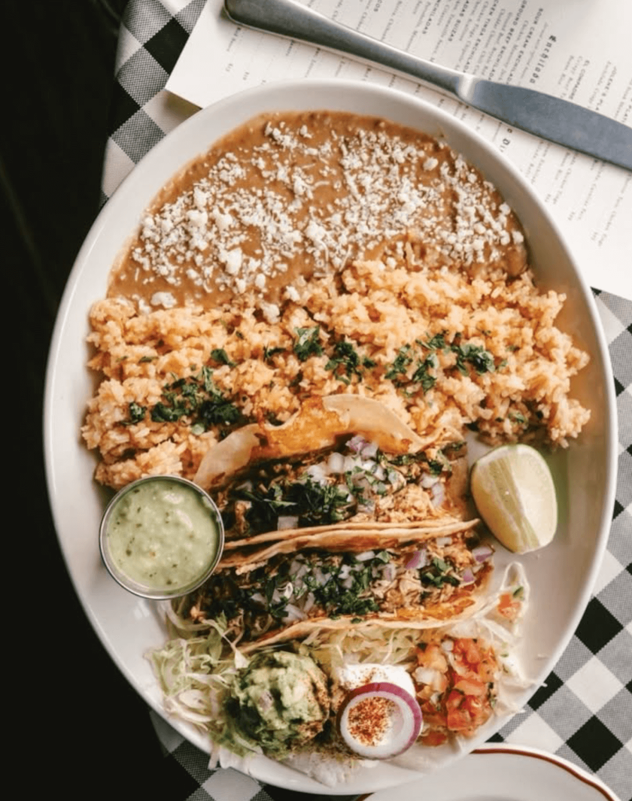 A plate with refried beans, Mexican rice, three tacos topped with onions and cilantro, guacamole, salsa, shredded lettuce, a lime wedge, and a cup of green sauce.