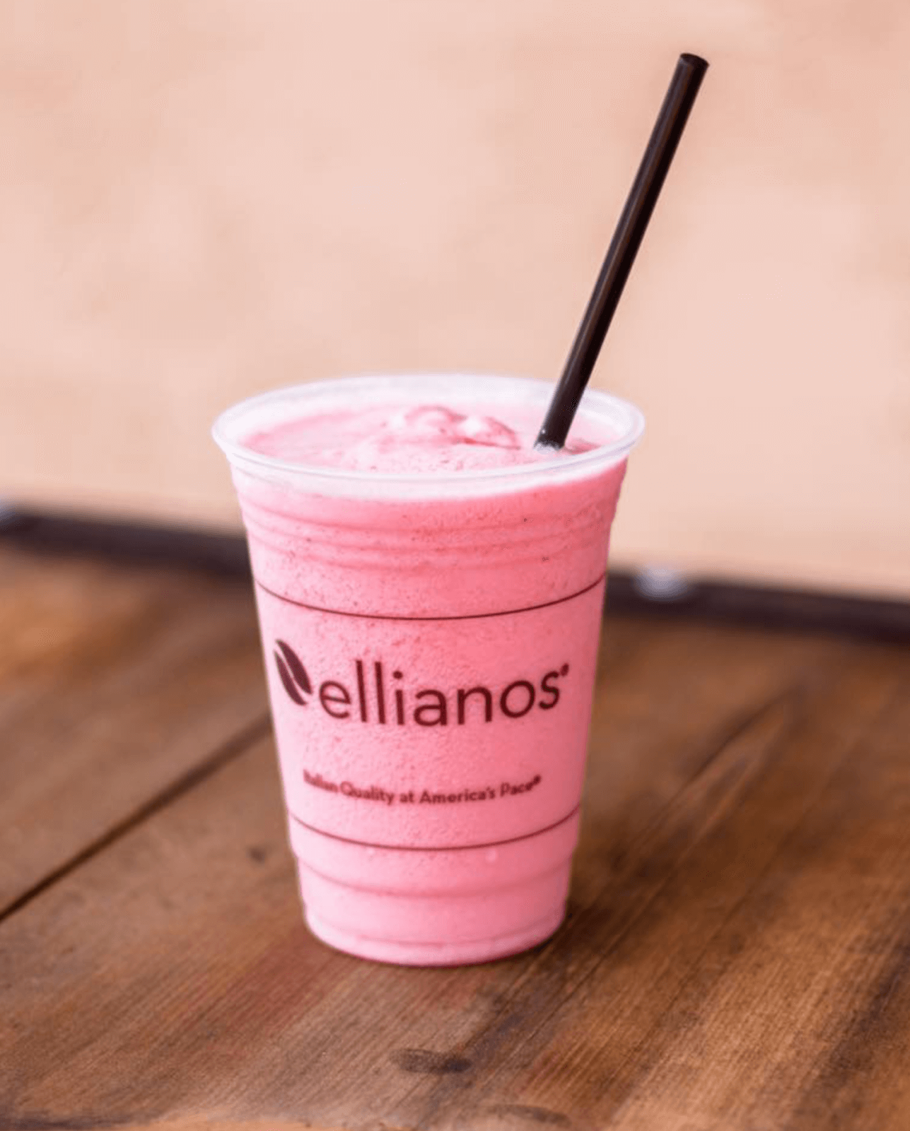 A pink smoothie with a black straw in a clear plastic Ellianos cup sits on a wooden surface, perfect for trying at new restaurants or exploring the latest Birmingham restaurants.