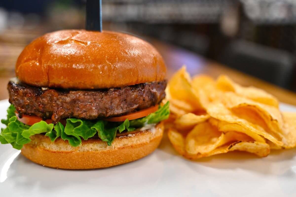 A hamburger with lettuce, tomato, and a beef patty in a brioche bun, served with potato chips on a white plate—perfect for exploring new restaurants in Birmingham or adding to your list of restaurants to try.