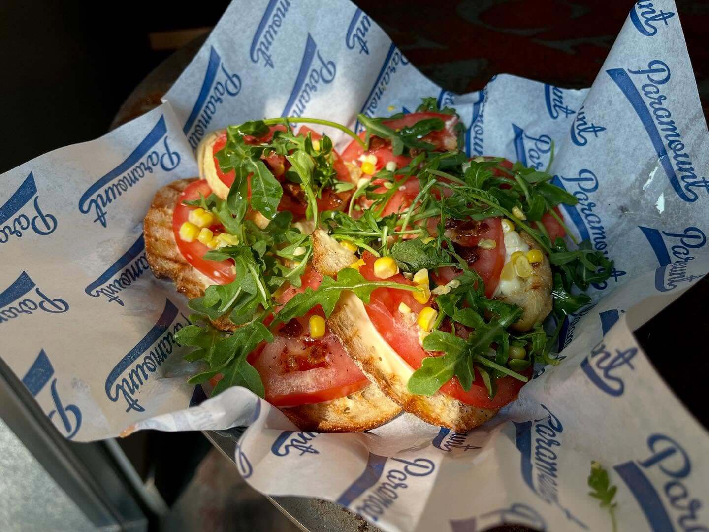 A serving of flatbread topped with tomato slices, arugula, corn, and herbs on Paramount-branded paper.