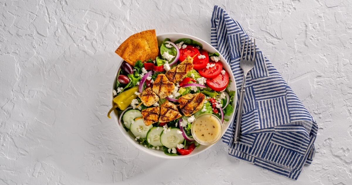 A bowl of Greek salad with grilled chicken, tomatoes, cucumbers, red onions, olives, feta cheese, pepperoncini, and pita chips sits beside a striped napkin.