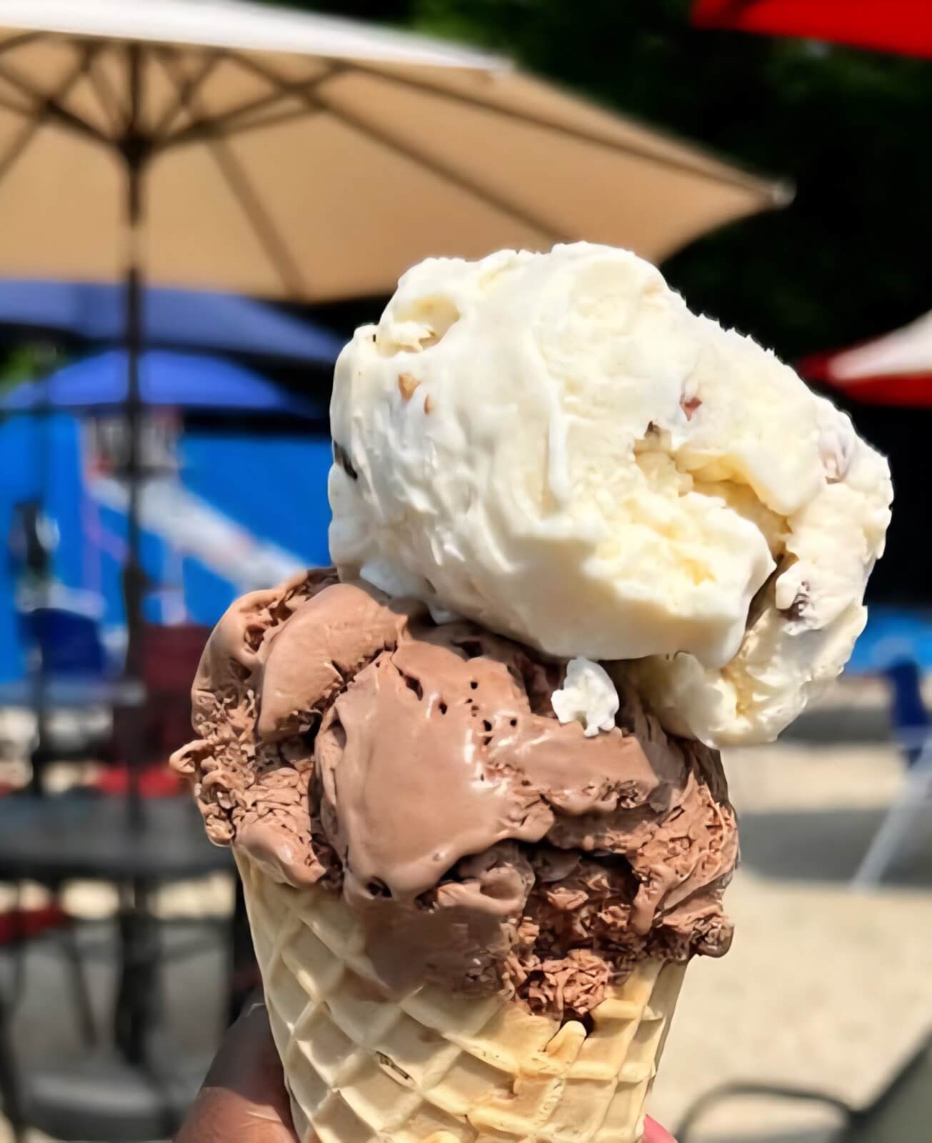 A hand holds a waffle cone with one scoop of chocolate ice cream and one scoop of vanilla ice cream outdoors at one of the new restaurants Birmingham has to offer, with umbrellas and tables in the blurred background.