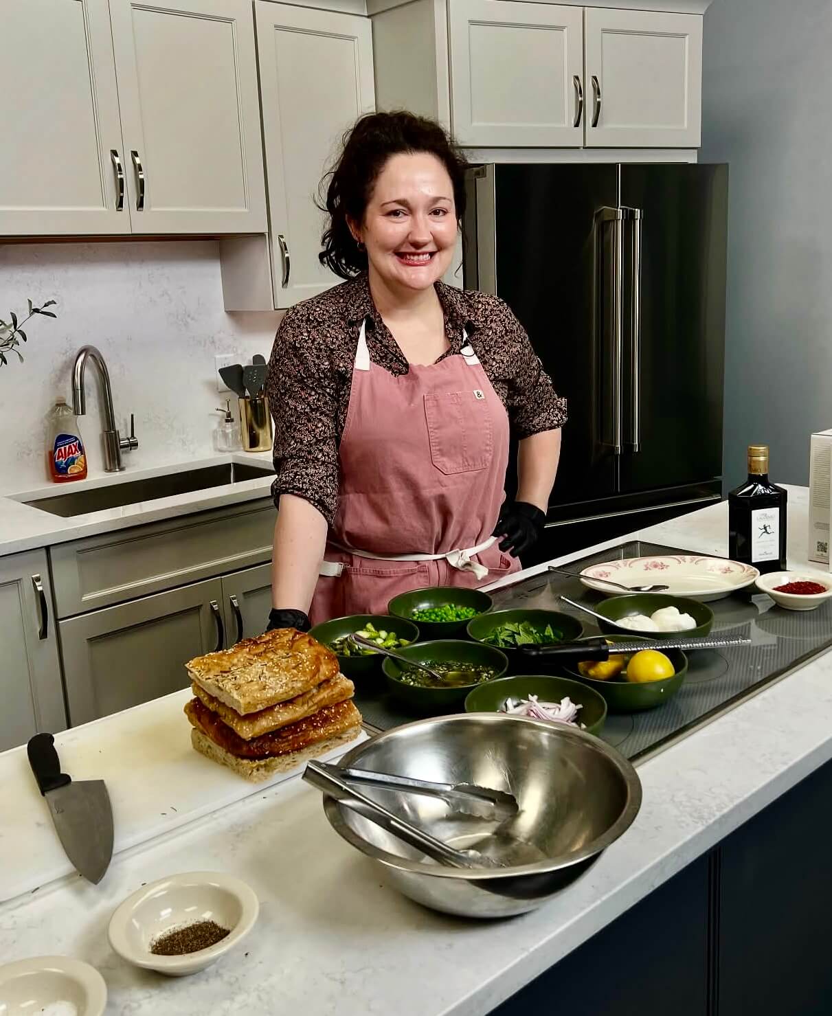This BHAM Chef Brings Heart & Heritage to Homewood - 1 Chef Rita Bernhardt, wearing a pink apron, stands in a Homewood kitchen behind a counter with various ingredients, bowls, and utensils prepared for cooking.