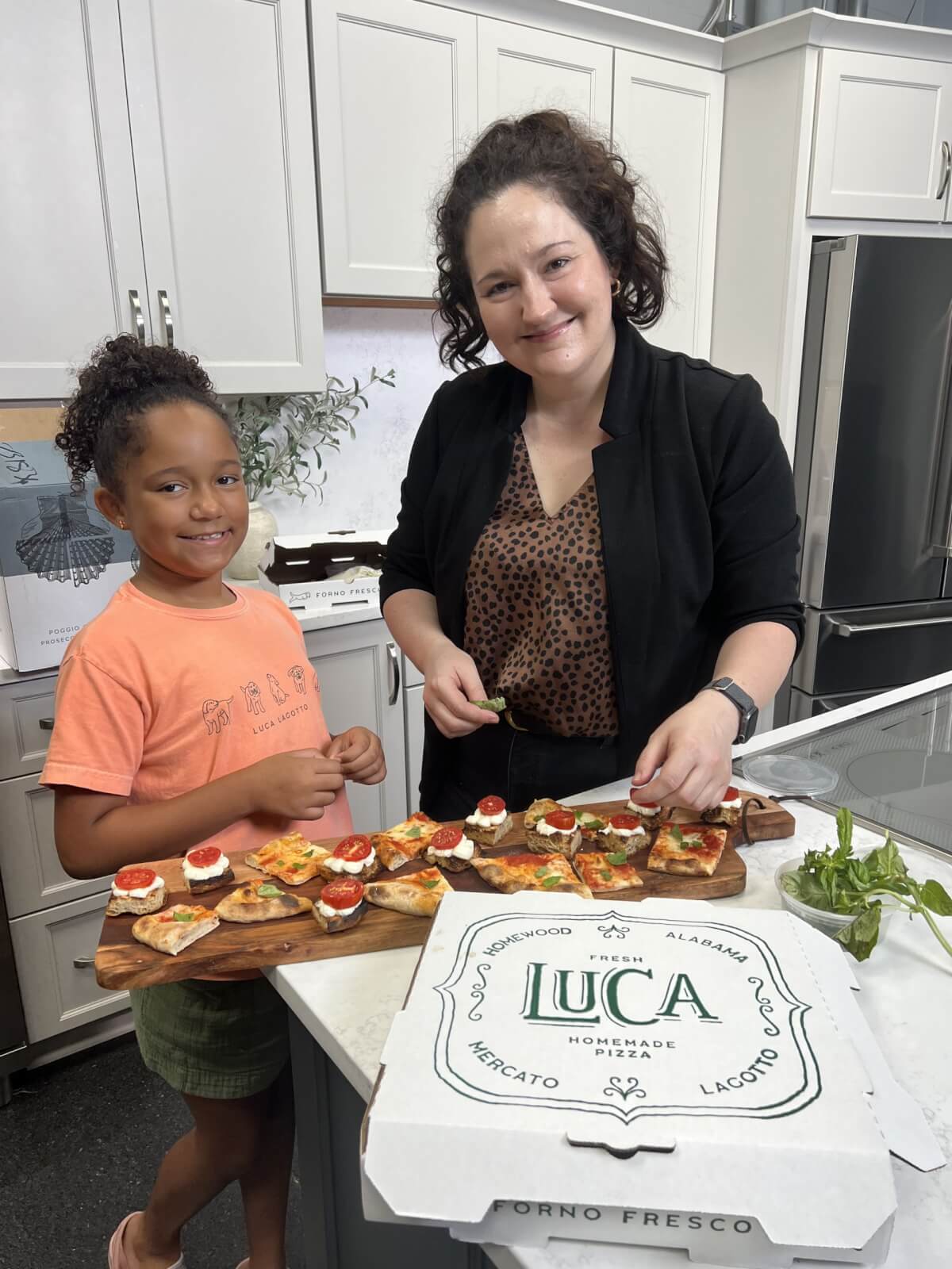 This BHAM Chef Brings Heart & Heritage to Homewood - 2 An adult and a child stand in a Homewood kitchen preparing appetizers on a wooden board, with a pizza box labeled "LUCA" and fresh basil on the counter.