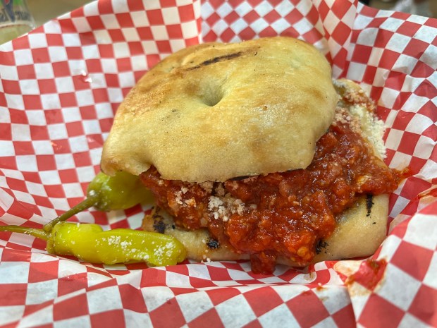 A focaccia bun filled with bright red meat and marinara filling. There are pepperoncinis on the side