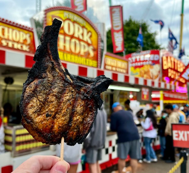 A charred pork chop on a stick is held up in front of a colorful Fair stand