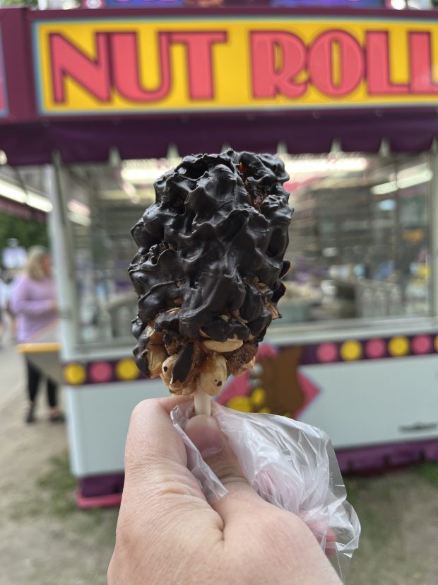 A dark chocolate covered nut roll on a stick is held up in front of a sign that reads NUT ROLL