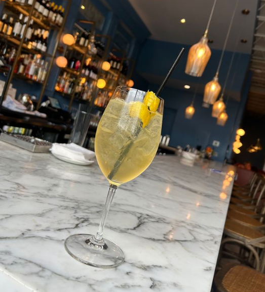 A angled shot of a wine glass with effervescent liquid and a lemon rind. Bar in background