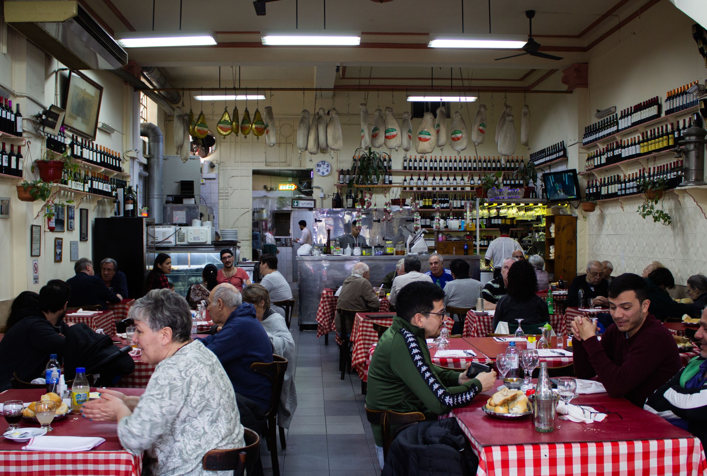 A packed restaurant interior with diners at red checkered tablecloths, hunks of meat hanging from the ceiling, cooks working behind a counter in the back, and rows of wine bottles on the walls.