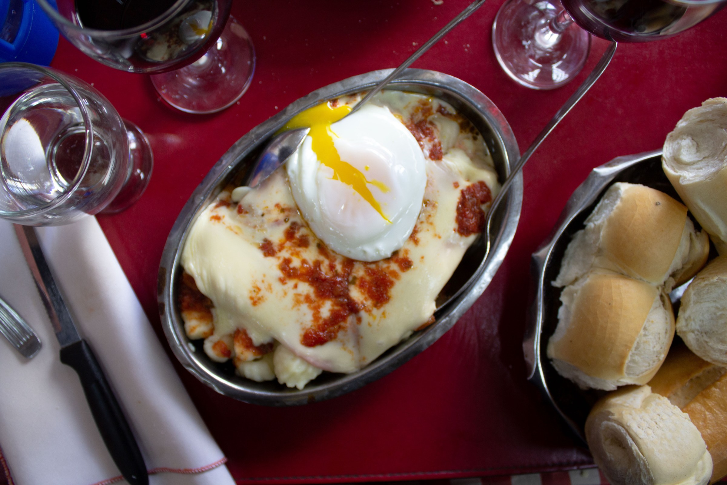 From above, a metal dish with tongs. The ñoquis are invisible beneath a blanket of cheese and a runny egg.