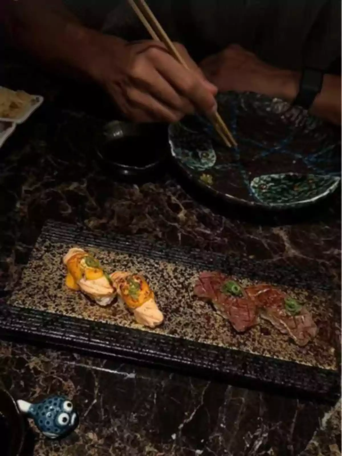 Person using chopsticks to eat sushi in an upscale restaurant with a marble table and decorative plates