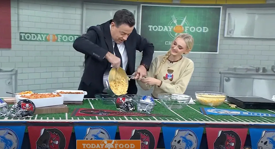 Two people at a TV cooking show table prepare a dish with ingredients and utensils, surrounded by football-themed decor from various teams