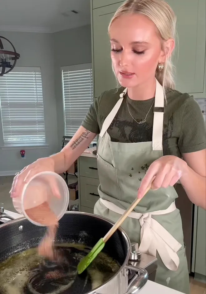 Person cooking in a kitchen, wearing a green shirt and apron, stirring a pot on the stove while pouring in an ingredient from a small bowl