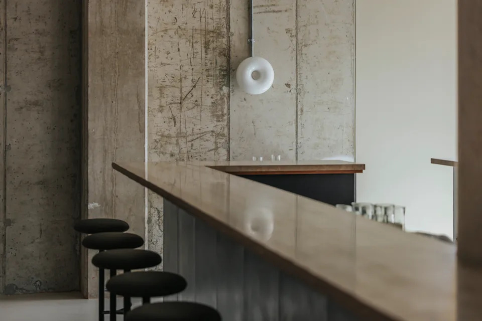 A restaurant bar counter pairs matte metallic surfaces with tall minimalist black stools, a wooden slab, and exposed cement walls adorned with a doughnut-shaped lamp.