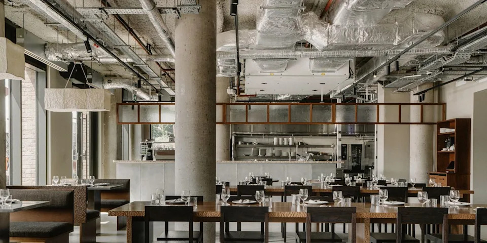  A brutalist restaurant with exposed cement walls, silver ventilation pipes, wooden furniture, and chrome-clad decor is ready for lunch service and brought to life by light. 