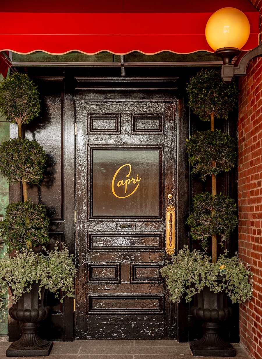 The shiny black door to a restaurant has a tree on either side of it and the word Capri in gold cursive on it.