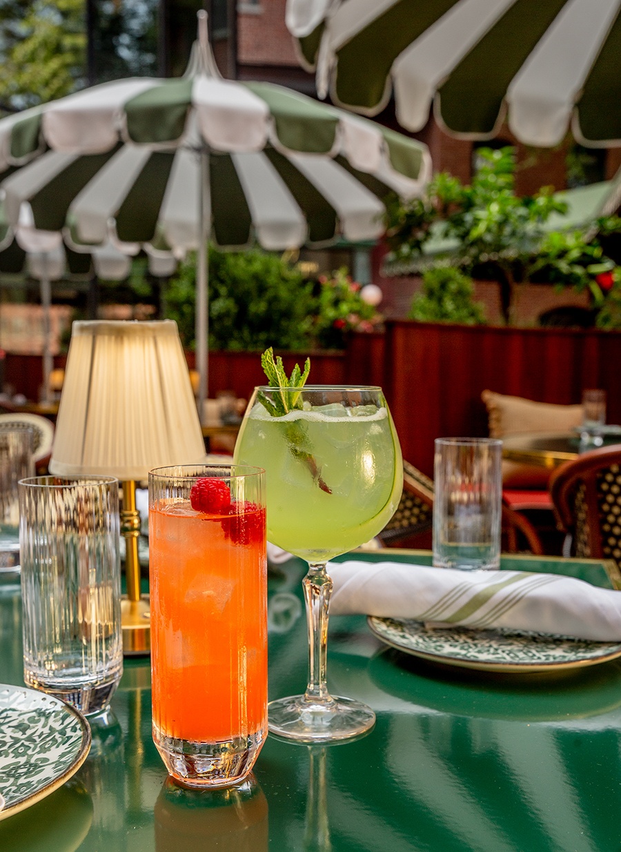 Summery cocktails sit on a green table on a restaurant patio with green-and-white-striped umbrellas visible in the background.