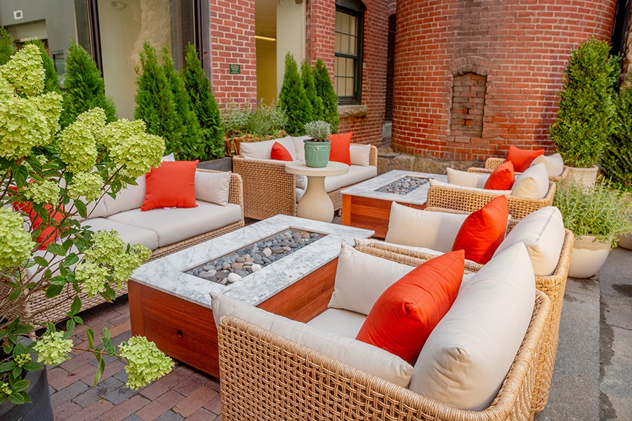 Beige and orange lounge seating around fire pits on a restaurant patio with lots of red brick in the background.