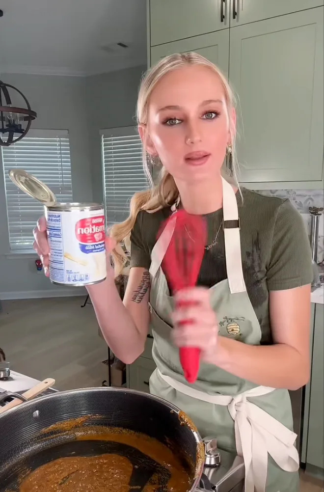 Person cooking in kitchen, holding a can of evaporated milk, wearing an apron, whisking a mixture in a pot