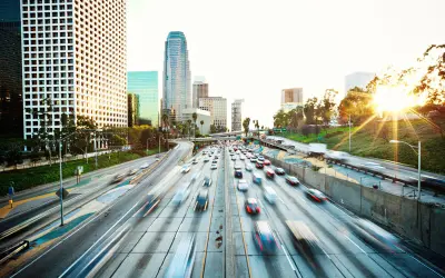 An urban freeway with fast-moving traffic, captured with motion blur to emphasize speed. Surrounding the freeway are modern skyscrapers and lush greenery. The setting sun casts a warm glow over the scene, with bright sunlight peeking through trees on the right.