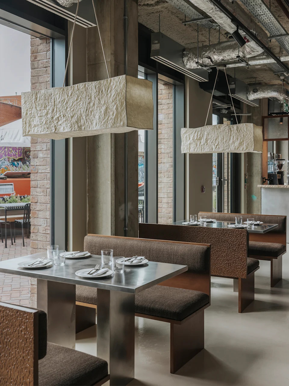A restaurant with brutalist interiors features wooden upholstered banquettes with warm, brown fabric matching with matte chrome tables with a geometric shape.