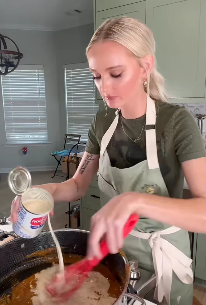 Person cooking in a kitchen, pouring evaporated milk into a saucepan while stirring the contents with a spatula
