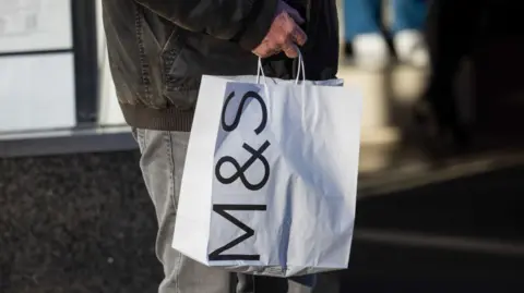 Getty Images Man in dark winter jacket and grey jeans carries white paper bag with M&S logo on the side 