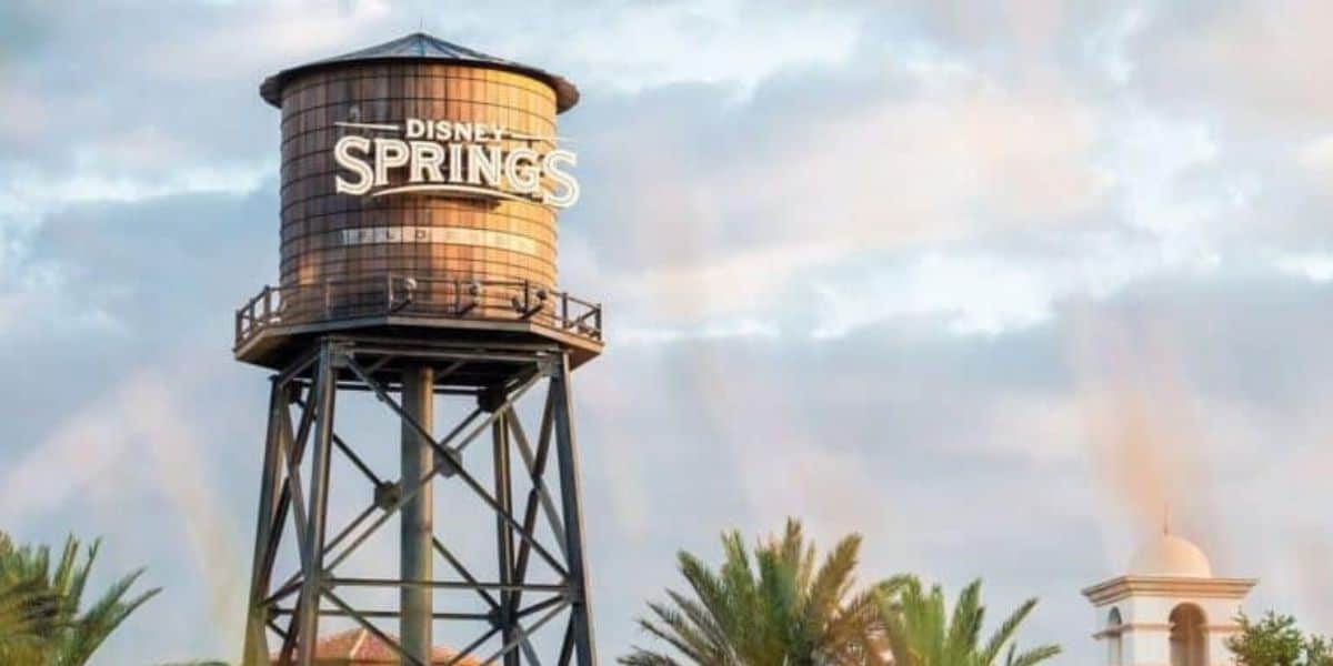 A large water tower with the words "Disney Springs" painted on it stands against a cloudy sky. Elevated on a metal frame and surrounded by palm trees and nearby buildings, the scene is lit by soft, warm evening light, evoking the enchanting atmosphere of Disney World at Disney Springs.