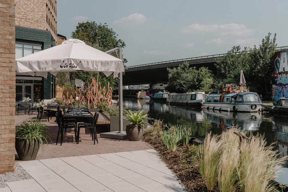 A sun-lit restaurant features brutalist interiors paired with a minimalist, black wrought iron-furnished canal-view terrace.