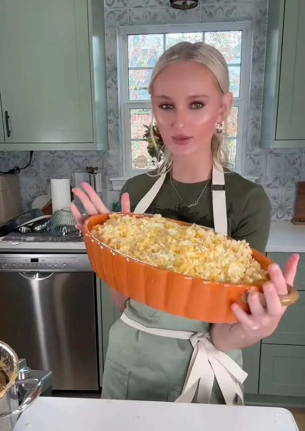 Person in a kitchen holding a large oval dish of prepared pasta casserole, wearing a t-shirt and apron