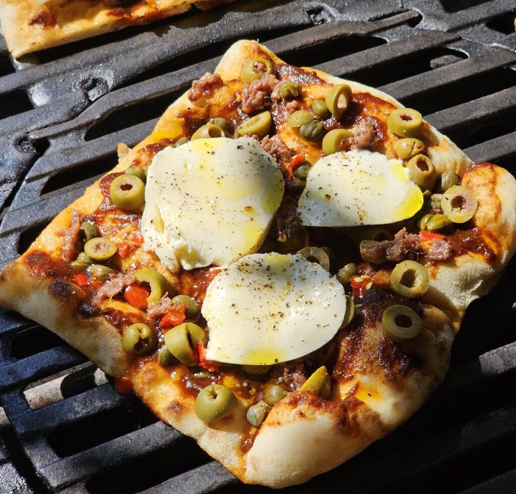 Pinsa fresh from the Grill (with tomato sauce from last harvest, capers, olives, anchovie fillets, chilies, mozzarella, olive oil, and black pepper), alongside a "Gschprützte Wiisse"