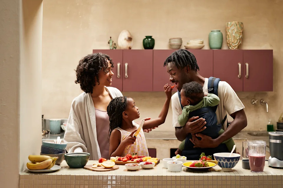 Girl feeding strawberry slice to father carrying toddler in carrier by mother at illuminated kitchen