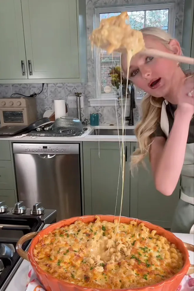 A person in a kitchen lifts a portion of cheesy baked macaroni from a large dish, showcasing its stretchy cheese