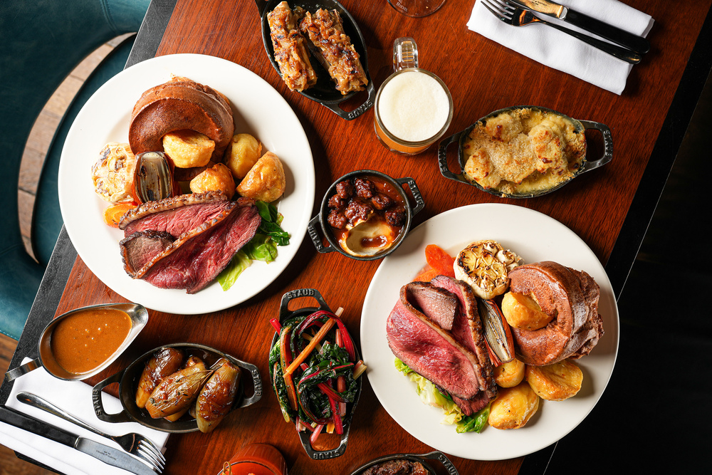 two sunday roasts platters piled high with rare beef and plnty of veggies, alongisde sides like cauliflower cheese, buttered veggies, and more