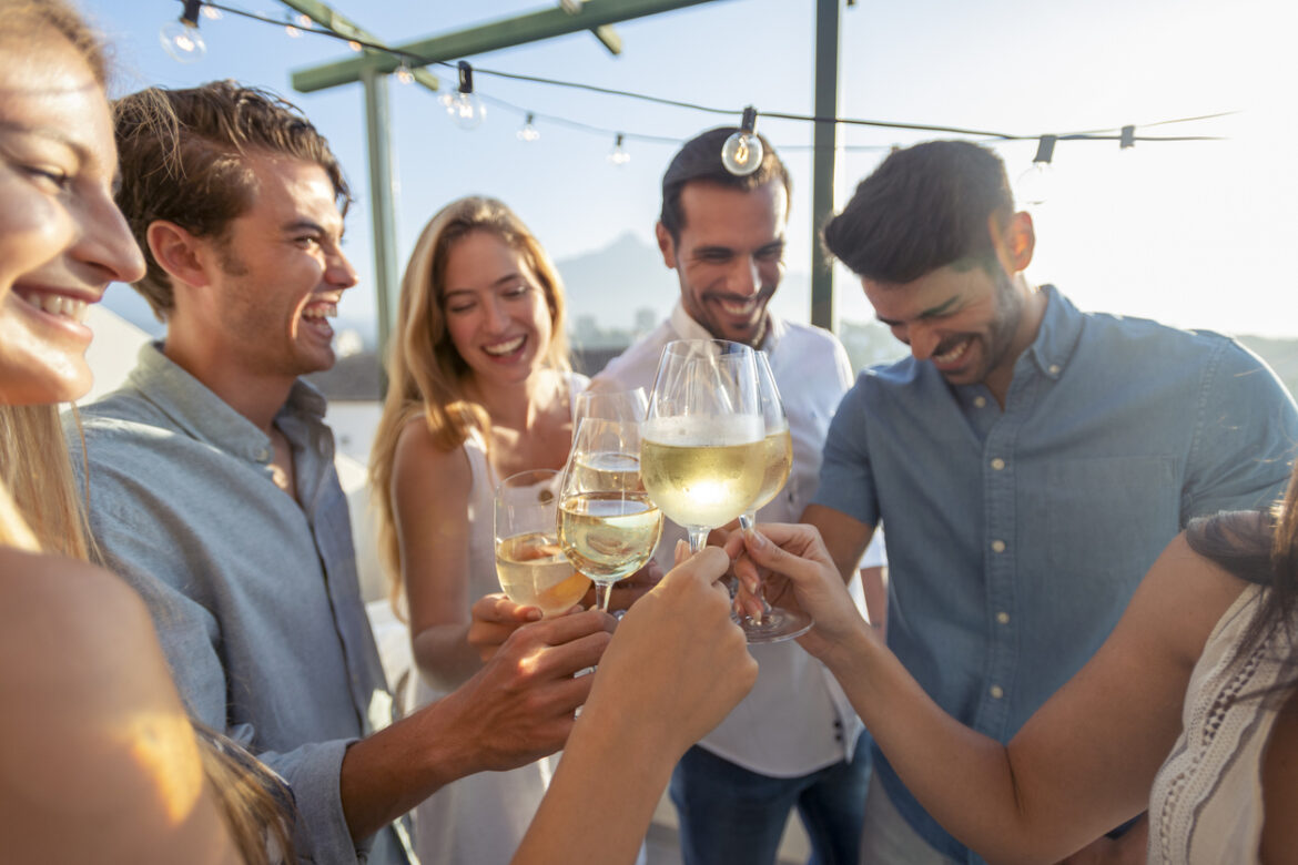 Group of friends having drinks at sunset. They are celebrating with a wine toast. They are drinking white wine. They are smiling, laughing and having fun.