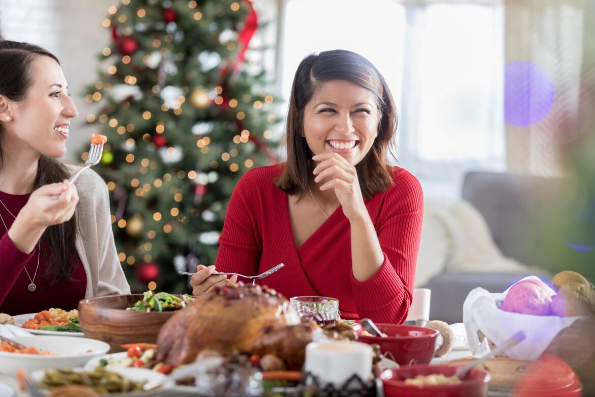 Two friends are eating at a table during a Christmas dinner celebration They laugh and smile together with other unseen friends.