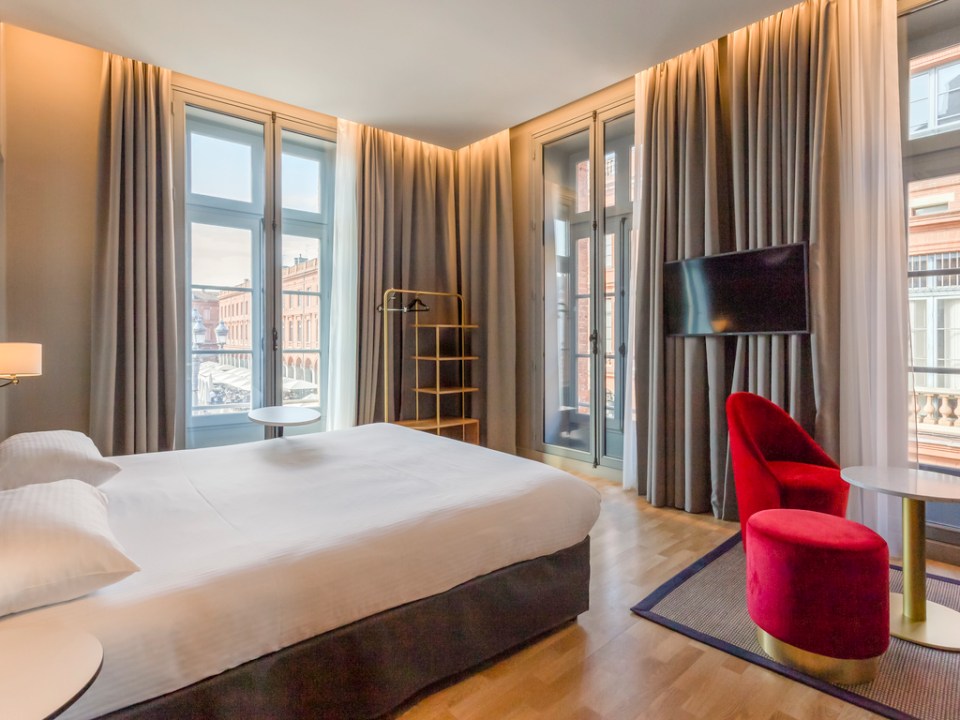 Hotel room with a bed, red chair and ottoman, and city view.