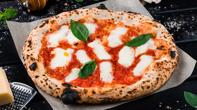 Pizza on culinary paper against dark background surrounded by ingredients and cooking tools