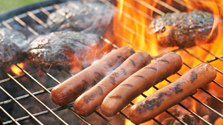 hot dogs on a flaming grill with burgers in the background