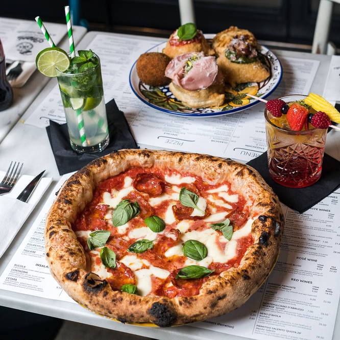 a pizza presented on a table next to two cocktails and a platter of appetizers and small bites