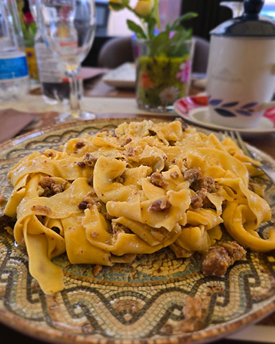 Plate of tagliatelle pasta with creamy sauce and pieces of meat, served on a decorative mosaic-patterned plate with glasses, flowers, and tableware in the background.