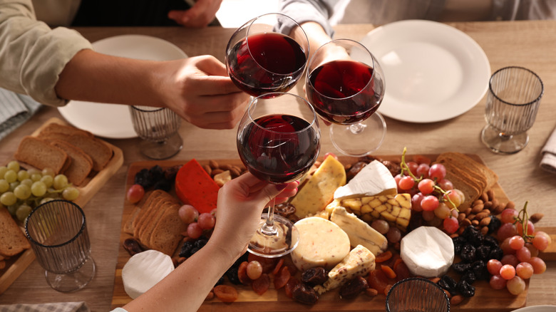 Three hands toasting red wine over cheese board