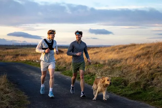 Medium shot of two young adult males and a dog running along a country lane in Northumberland, North East England.