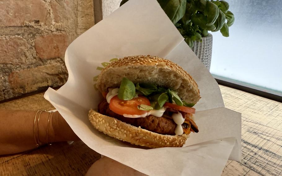 A sesame seed bagel sandwich filled with a breaded veggie cutlet, fresh tomato slices, lettuce, and white sauce, wrapped in white paper and sitting on a wooden table next to a small potted basil plant by a window.