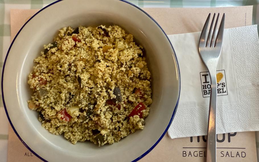 A bowl of couscous salad mixed with diced vegetables including red peppers and herbs, served in a white bowl with blue rim on a striped placemat with a fork alongside.