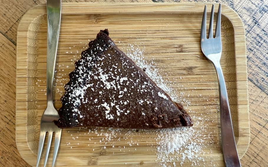 A triangular slice of rich chocolate cake dusted with powdered sugar, served on a wooden cutting board with a knife and fork on either side.